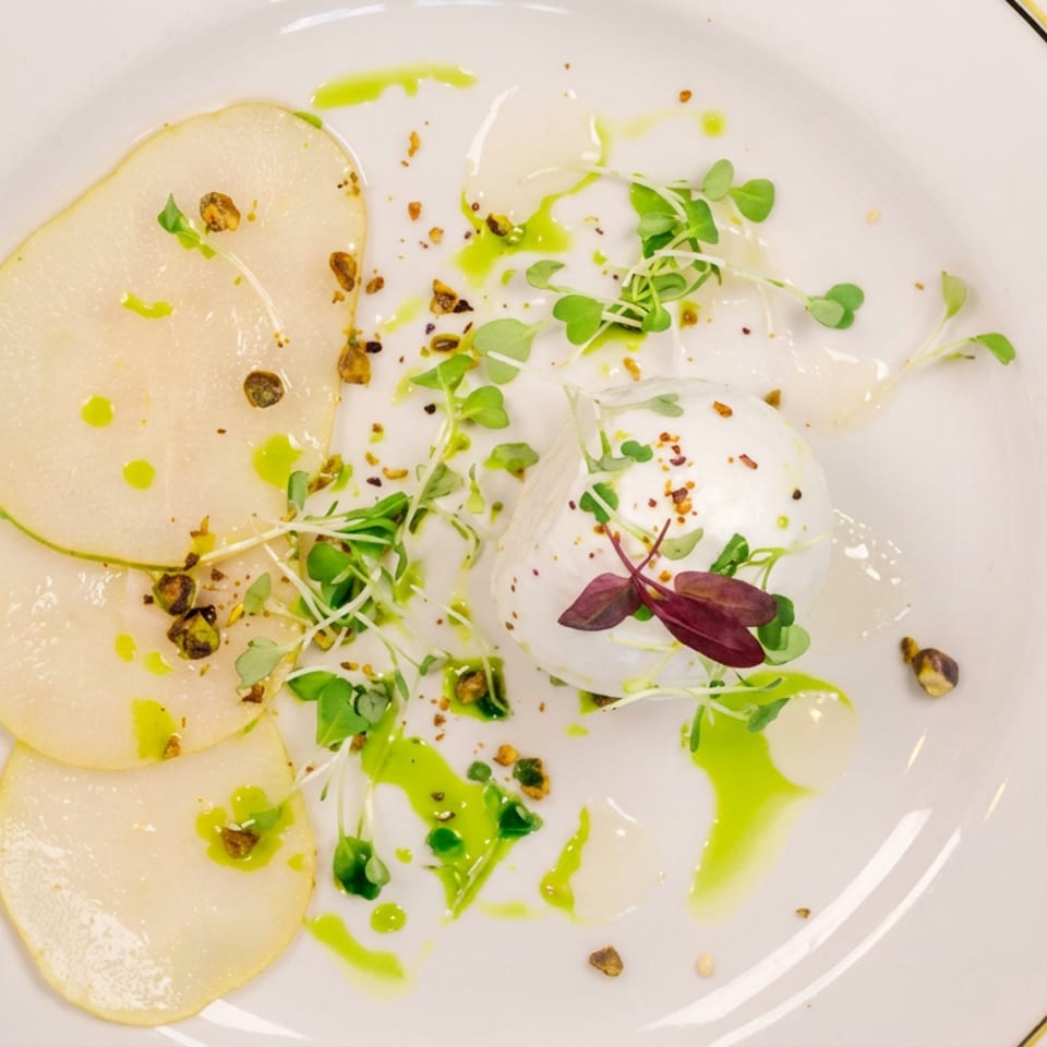 Elegant plated dish with thin pear slices, creamy white elements, microgreens, pistachios, and bright green oil on a white plate.