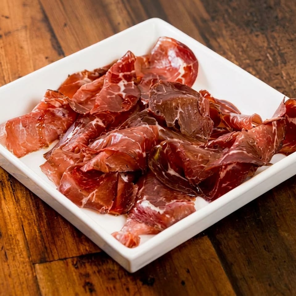 Thinly sliced cured meat arranged on a white square plate atop a rustic wooden table.