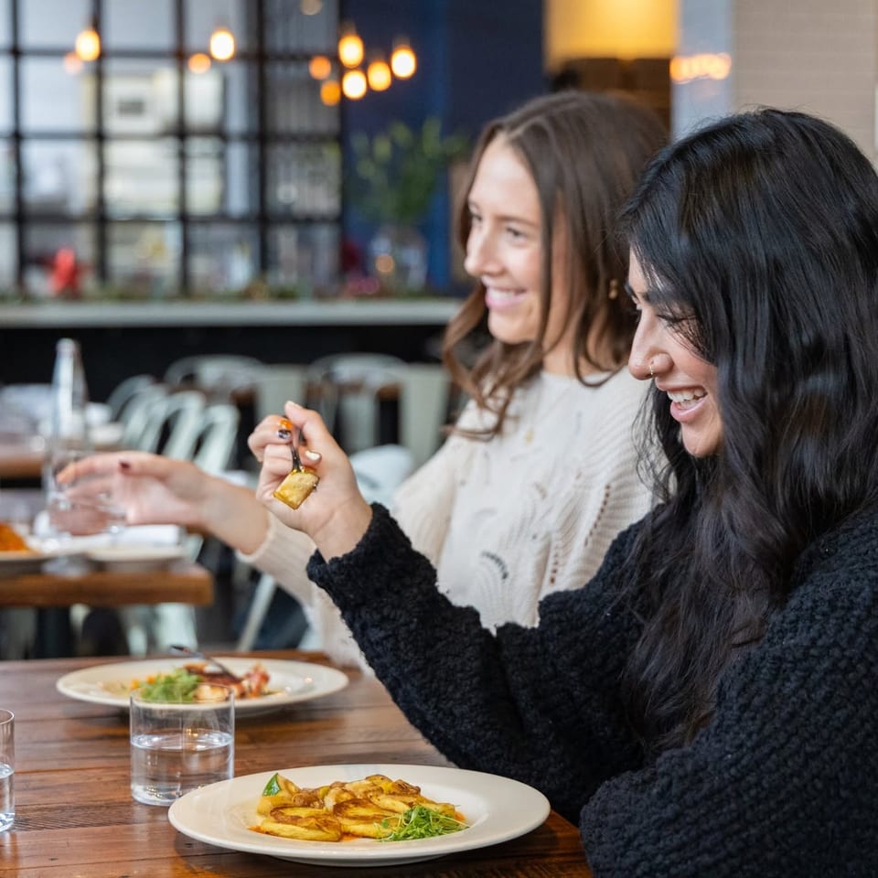 Two women laugh over plated meals at a warmly lit restaurant table.