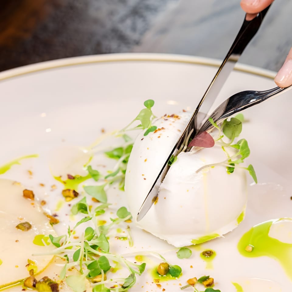 Fine dining dessert being cut with a knife and fork, garnished with microgreens, pistachios, and green sauce on a white plate.