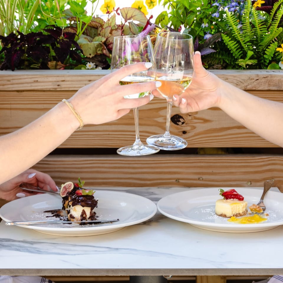 Two diners toast with white wine over chocolate and cheesecake desserts at a wooden restaurant table surrounded by colorful plants.