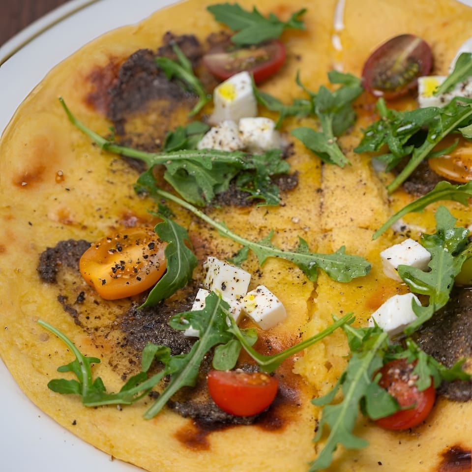 Close-up of a golden flatbread topped with arugula, cherry tomatoes, feta cheese, herbs, and black pepper.