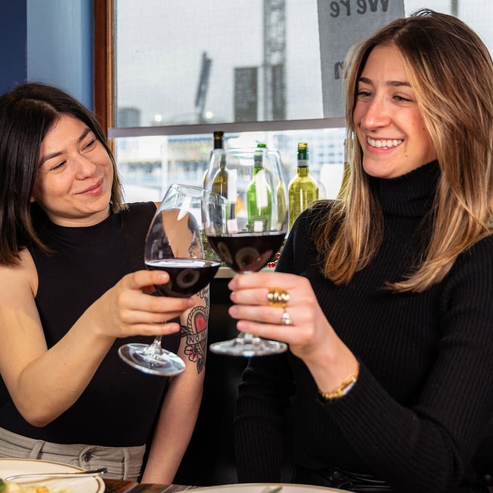Two smiling women toast with glasses of red wine at a restaurant table beside a window.