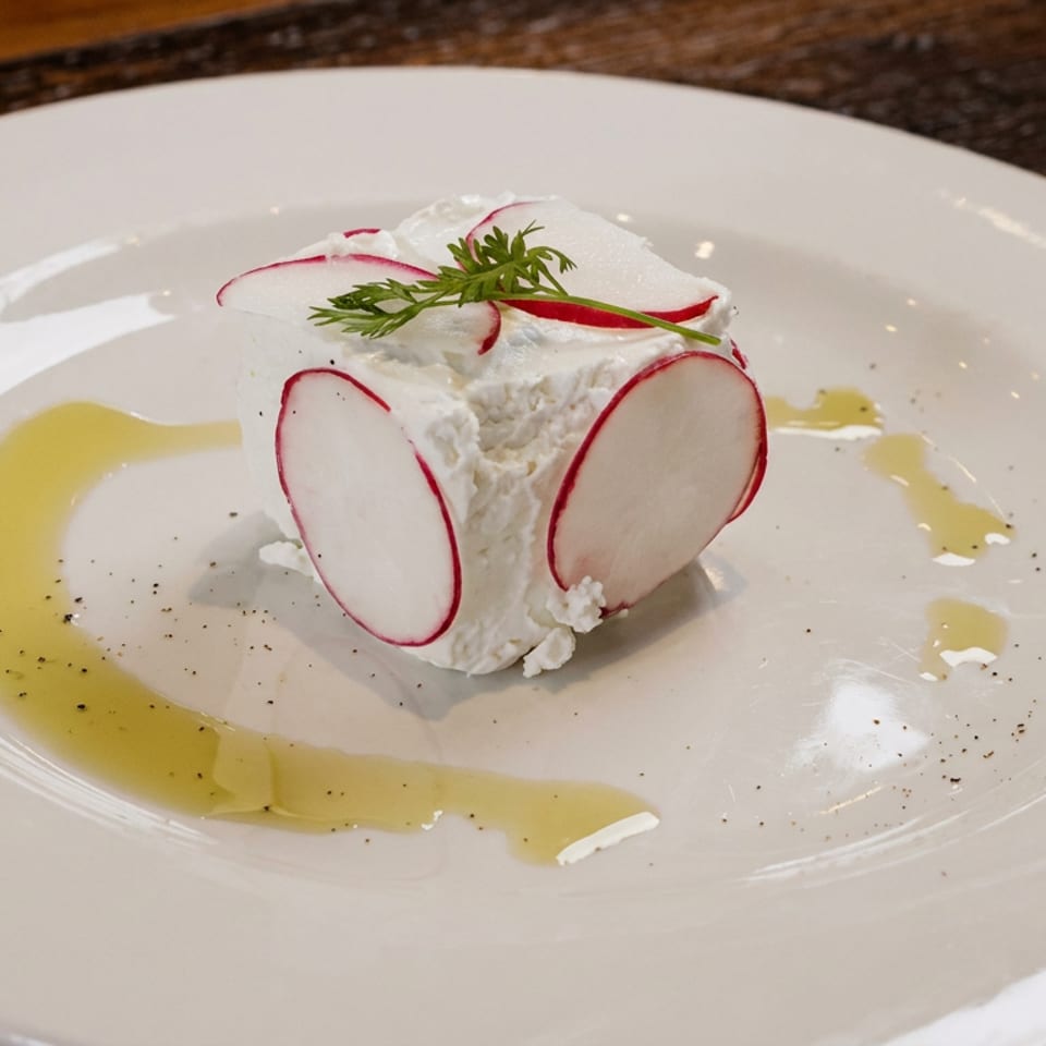 Creamy white cheese topped with radish slices and herbs, surrounded by olive oil and cracked pepper on a white plate.