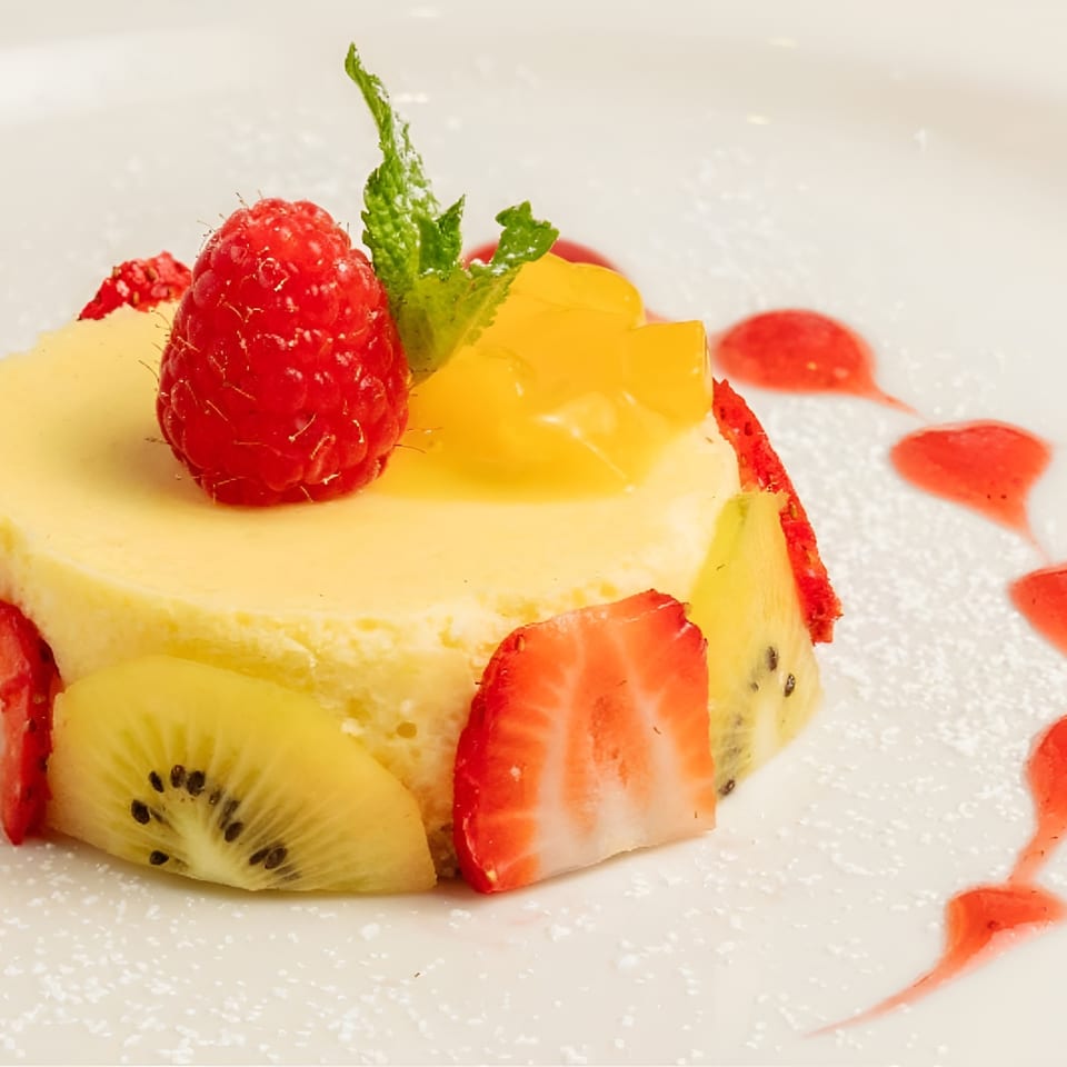 Fruit-topped cheesecake garnished with raspberries, strawberries, kiwi, mango, and mint on a white plate with raspberry sauce.