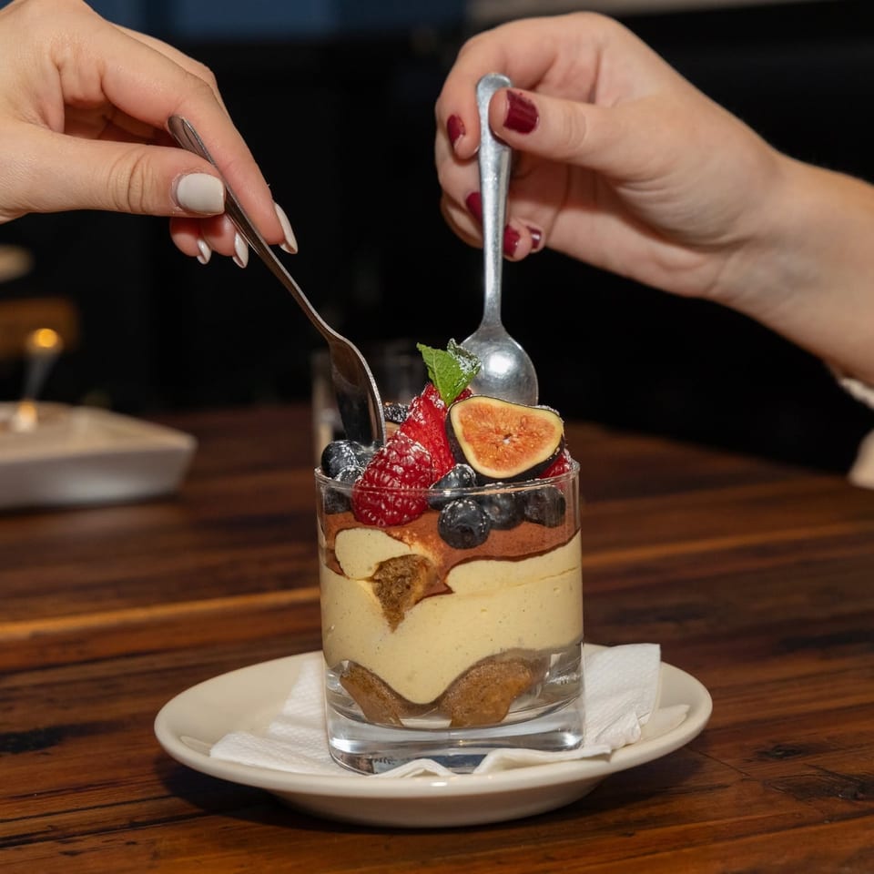 Layered dessert topped with raspberries, blueberries, sliced fig, mint, and powdered sugar, served in a glass on a restaurant table.
